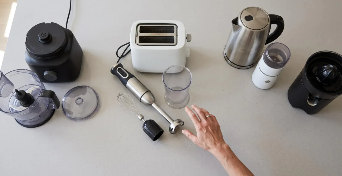 Vue du dessus de plusieurs petits électroménagers de cuisine disposés par catégories sur un plan de travail avec une main sélectionnant un appareil